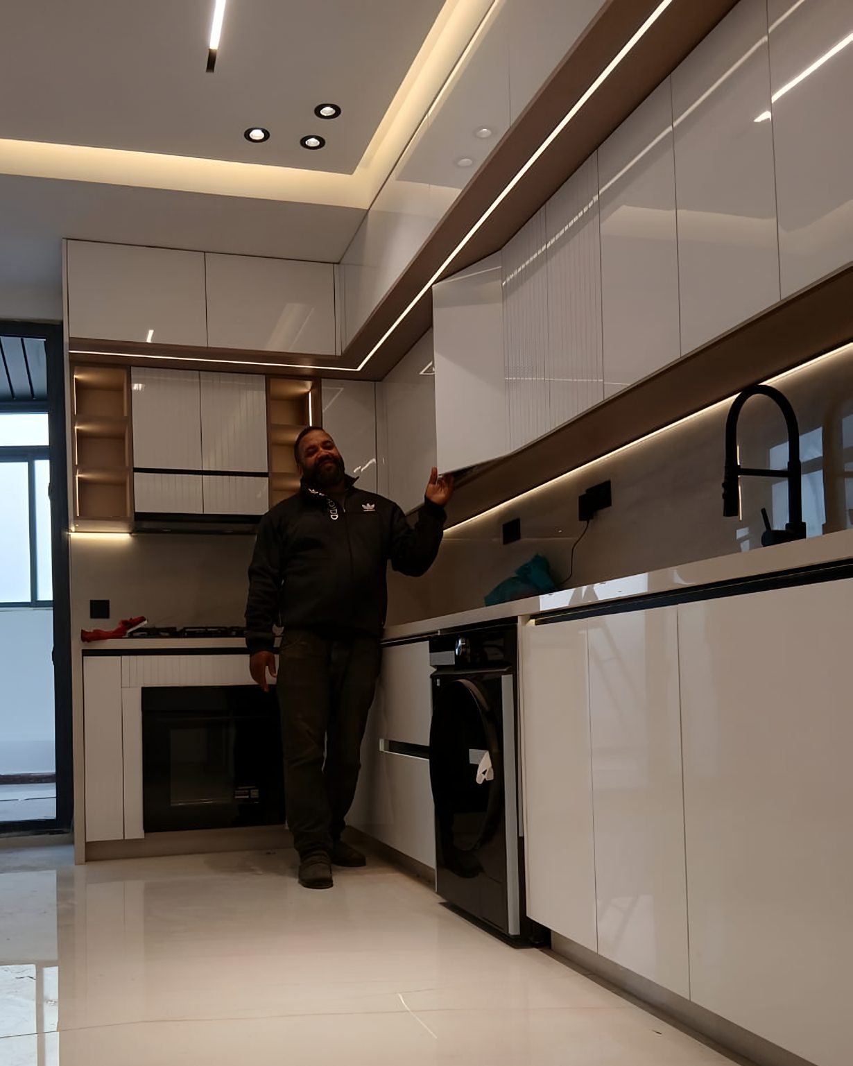 Cuisine blanche aux façades brillantes et cannelées, éclairage intégré au plafond et sous les meubles hauts, un homme debout au milieu de la pièce